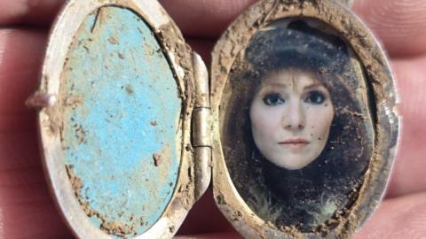 An open silver locket, covered in mud, containing the image of a 20th century woman, wearing eyeliner and a cap. The photograph looks to be taken in passport-style.