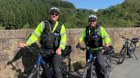 Two police officers on bikes by a wall with trees in the background and blue skies