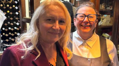 Two women look at the camera. The woman on the left has long blonde wavy hair and wears a wine-coloured suit jacket with a black top. The woman on the left has dark hair and glasses and is wearing a white collared shirt and a dark pinafore dress. Wine bottles are in the background.