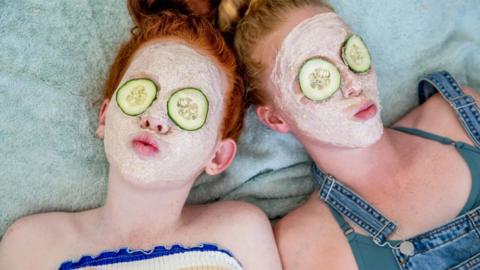 Two young girls lying on a towel with a cream face mask on, slices of cucumbers over their eyes