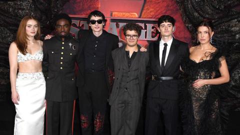 The cast of Stranger Things stand in mostly dark clothing in front of the show's branding at the show's Dubai premiere. Sadie Sink, Caleb McLaughlin, Finn Wolfhard, Gaten Matarazzo, Noah Schnapp and Millie Bobby Brown are all in attendance.
