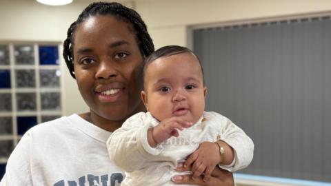 Sharna holds a child. She is in a white jumper and the child is in a cream-coloured baby suit with the words 'little one' embroidered onto it.