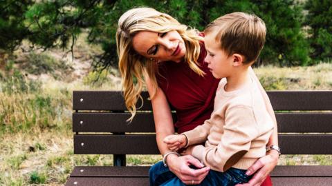 A mother talks to a child sitting on her lap on a park bench