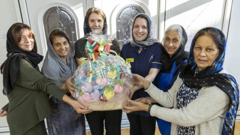 Six women, all wearing head scarves, have their arms outstretched in front of them and are holding a basket filled with colourful knitted hearts. The basket is wrapped in plastic and tied with a colourful ribbon