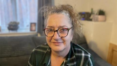 Sarah is sitting on a grey ribbed sofa and is wearing a green, black and grey checker shirt and wearing red black and brown turtle shell glasses. The background is blurred.