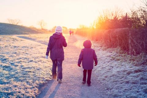 Stock photo of a woman and boy pictured from the back wearing coats and other winter clothes as they walk along a path in the snow in the sunshine.