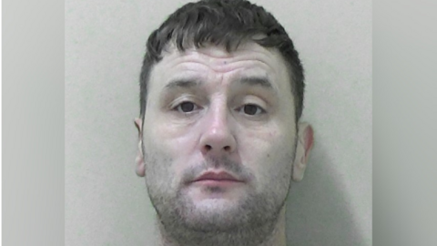 A police mugshot of Gary Rarity, a man in his 40s with stubble, dark eyes, and a dark fringe-style haircut with shaved back and sides.
