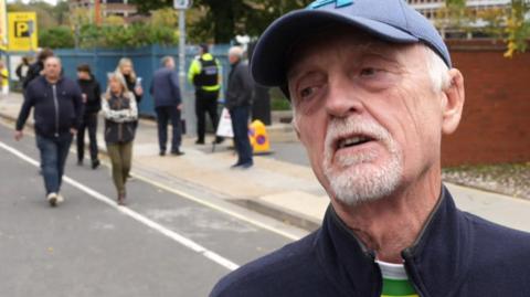 An older man with a white goatee beard, in a navy fleece, and with a blue baseball cap, talks to a reporter who is standing off camera. There are people walking on a road outside Portman Road stadium behind him.