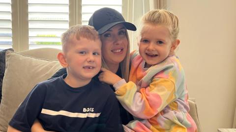 Ashley, who is wearing a black baseball cap and black T-shirt, is sitting on the couch in her home in front of a window with her two children. They are all smiling at the camera.