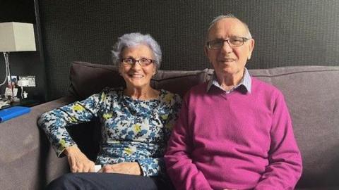 An elderly couple sit on a sofa smiling at the camera , She wears a flowery top, he wears a purple jumper.