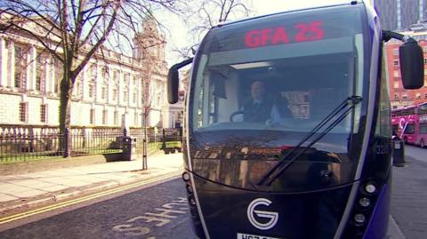 A purple-and-black trackless tram, known as a "glider bus", is about to pull away from the kerb in Belfast city centre.