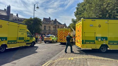 Several yellow‑green NHS ambulances and an incident response unit are parked on a street, with an ambulance worker in uniform at the rear of one vehicle and people standing nearby beside historic stone buildings under a clear sky.