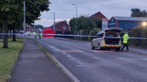 A blue and white police cordon is stretched across Cottage Beck Road in Scunthorpe, as officers in yellow high-visibility jackets investigate the scene of a crash. In the background is a large red tent.