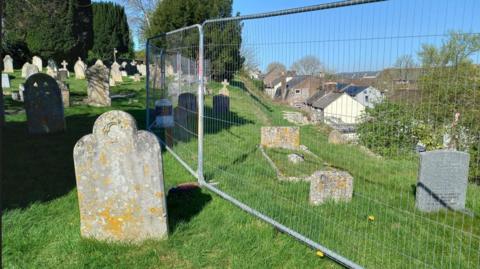 An area of Fordington Cemetery above the wall collapse. There are barriers around some of the graves. It's a sunny day.
