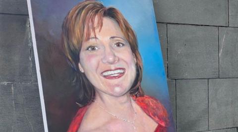 A portrait of a woman with brown hair, wearing red, is by a wall.