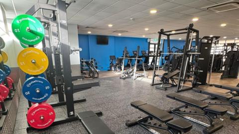 The inside of a gym with weights stacked up on the left hand side and weightlifting machines lined up on the right.