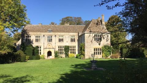 The frontage of a small country house on a sunny day. A lawn and armillary sphere can be seen in the foreground, with trees in the background.