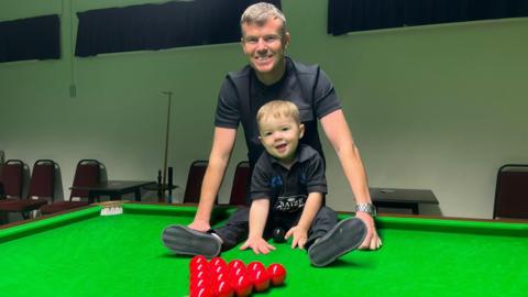 Jude is sitting on top of the snooker table with the balls in front him. His father Luke is standing behind him. They are both smiling at the camera. 