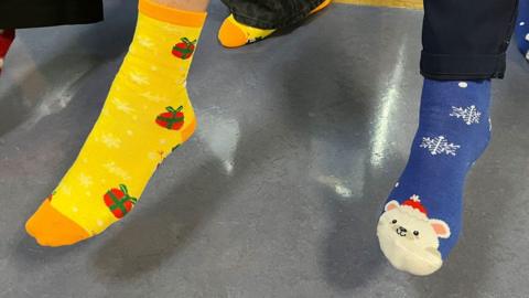 A close up of two right feet, one clad in a bright yellow sock with a red and green parcel on it and the other in a blue sock with a red hat wearing polar bear and white snowflakes.