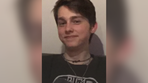 A teenage boy with brown hair styled with a fringe on the left covering part of his forehead looking into the camera. He is wearing a black t-shirt with a white logo, a small silver coloured chain and a black chain beneath it.