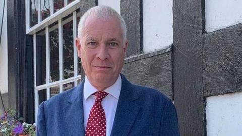 Councillor Ewen Sinclair stands outside a building with a flower bed behind him. He has grey hair and is wearing a blue suit with a jacket and tie.