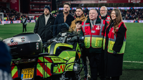 A row of Ian Coates' family and blood bike representatives standing behind a motorbike with fluorescent yellow livery and the word blood on the side.