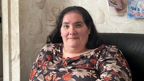 A woman in a floral dress sat on a leather chair and looking at the camera. She has a slight smile.