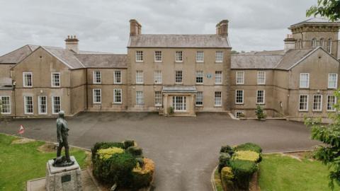 Royal School Dungannon. It is a large sandstone building with three stories in the main building and two stories the buildings connecting to it. The windows are large with white pain and lawns to the front with a statue on the left hand side.