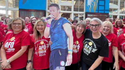 Female members of the All For One Choir stood in a large group inside Paragon Station. All of them are wearing red t-shirts with the words "Disneyland Paris: AFO Choir 2025" on them in white. One woman on the front row is wearing a black t-shirt. The are posing with a cardboard cut out of Chris Martin. 