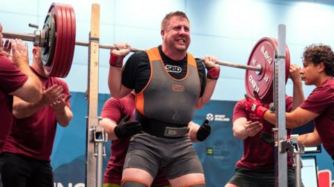 Chris Pryor wearing black shorts and a t-shirt underneath an orange and grey athletic vest and weight belt. He is straining as he squats a large weight, while spotters stand either side of him, ready to catch the barbell. 
