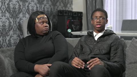 A woman and boy, both wearing black, sit on a grey sofa in a living room. A dining table and speakers can be seen behind them, as well as blinds. The woman has her forearms crossed and the boy has his hands in front of him, whilst speaking to the camera.