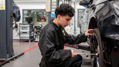 A student with curly black hair is squatted down next to a car. He is holding the car's tyre and is holding a metal piece of equipment that is attached to the valve of the tyre. He is wearing a fully black boiler suit with the college's logo in yellow on the breast. In the background is another car listed off the ground, and various garage equipment.