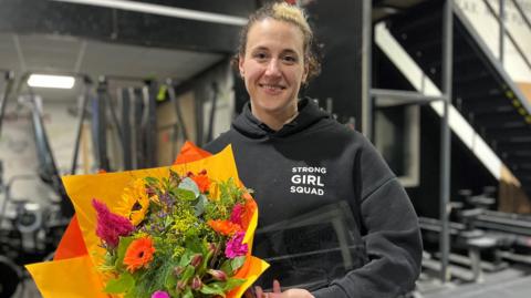 Kristen Ingraham-Morgan looks directly at the camera, smiling. She has brown eyes and brown hair, with a stripe of blonde. She is in a gym surrounded by equipment. Her black hoodie has 'Strong Girl Squad' printed in the top left-hand corner. She is holding a rectangular glass trophy with 'BBC Sports Personality Unsung Hero - East Yorkshire and Lincolnshire' written on it. In her right hand she holds a large bouquet of colourful flowers.