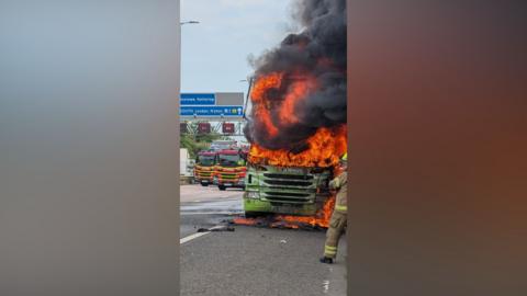 Two fire engines sit on the M1. The cab of a green lorry is on fire, with bright orange flames and black smoke.