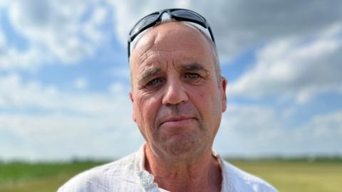 Peter Dixon has blue eyes, and has a pair of sunglasses on his head. He is stood in front of a field which has been blurred and a cloudy blue sky.