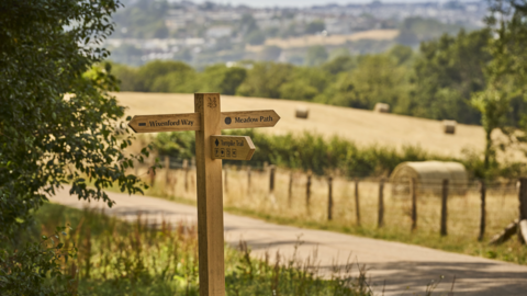 A wooden wayfinding sign stands beside a rural path bordered by grass, trees and fenced fields. The sign points to “Wixenford Way,” “Meadow Path,” and “Turnpike Trail.” In the background, hay bales dot a golden field, with trees and a distant town visible beyond.