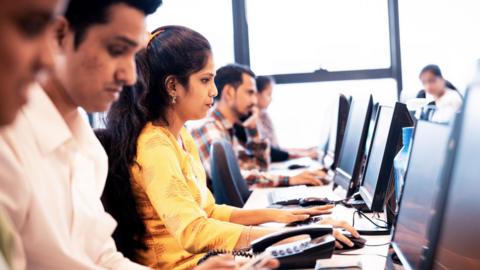The image shows Indian workers at a software company seated in front of their computer screens. A woman wearing a white kurta is prominently seen, surrounded by male colleagues on either side. 