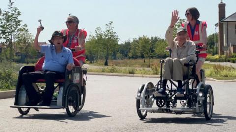 Two specially adapted trikes are being cycled, side-by-side, along a street. The trikes have two wheels at the front, wide enough apart to carry a passenger. They are powered and steered by women on the back, each wearing pink hi-viz vests. The passenger in the front of the trike on the left is an elderly man wearing a blue shirt and green baggy sun hat. He is holding a squeezy horn in the air. The passenger in the trike on the right is an elderly man wearing a tan cap - he is sat in a wheelchair which is mounted on the trike. He is smiling and waving.