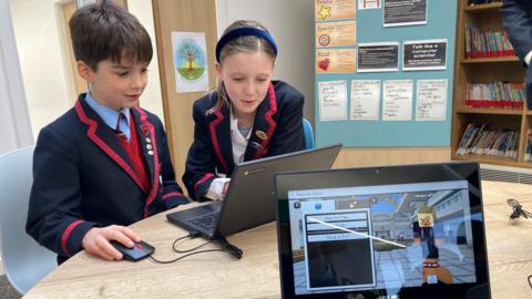 Two young children in red and black blazers, one boy and one girl, looking at black laptop on a round table. The boy is navigating a computer mouse. The background looks like a school with books on bookshelves and posters on the world. In the foreground - the camera is facing a laptop screen with the block-based world of Minecraft on the screen.