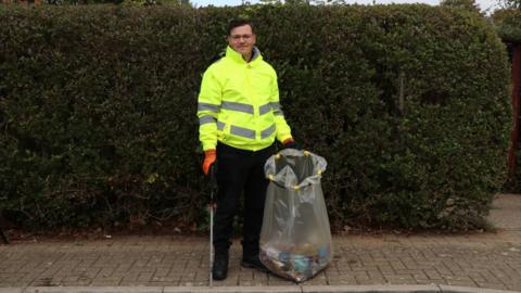 A man, Connor, stands on a pavement. He wears a hi-vis jacket and holds a litter picker and rubbish bag