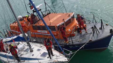 A large orange lifeboat pulled alongside a small white yacht. Crew members stand across both decks. The water is calm.