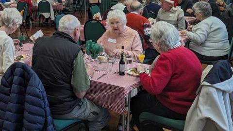 A group of elderly people sat around a table with glasses of wine and plates of food
