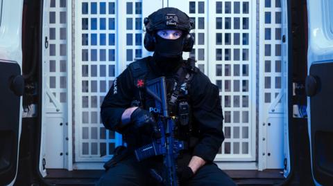 An armed police officer, holding his gun in a downward position and wearing a mask, looks toward the camera while sat in the back of a police van.