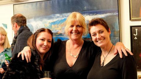 Three smiling women in black outfits stand in front of an abstract seascape painting.