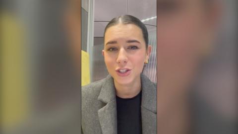 A woman with tied back brown hair wearing a grey blazer looks at the camera 