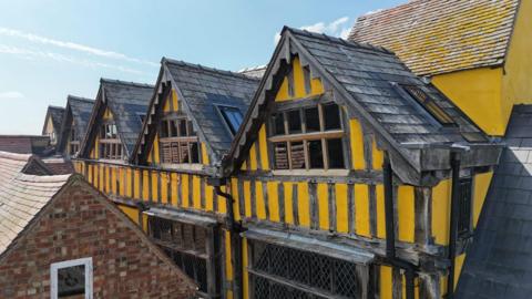 The restored building, it is yellow with black stripes, it has five adjacent gables with black tiled rooves.