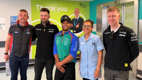 Four male race car drivers and one female nurse. The men are wearing mostly black apart from the one in the centre who has a green, blue and orange long sleeve top on. They are all smiling for a photo.
