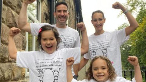 A family stand outside a house, wearing matching charity T-shirts that read "I've taken on a challenge for children with cancer".
