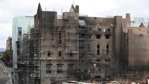 Large historic building undergoing restoration, surrounded by extensive metal scaffolding. The facade shows darkened, fire-damaged walls and empty window openings, while modern glass-sided structures are visible on either side. A wet street runs alongside the building, with two workers in high-visibility clothing visible in the lower left corner under an overcast sky