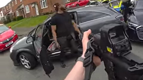 A man wearing a black T-shirt and black jeans stood next to a grey car with its door open as a gun points at him. Police cars can be seen behind his vehicle.